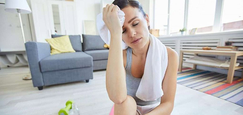 Young tired woman touching her forehead with towel while sitting on mat after hard workout due to iron depletion