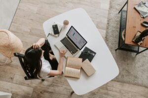 Overhead view of a woman working from home, sitting at a white table with a laptop, books, and coffee, illustrating a productive home office setup.
