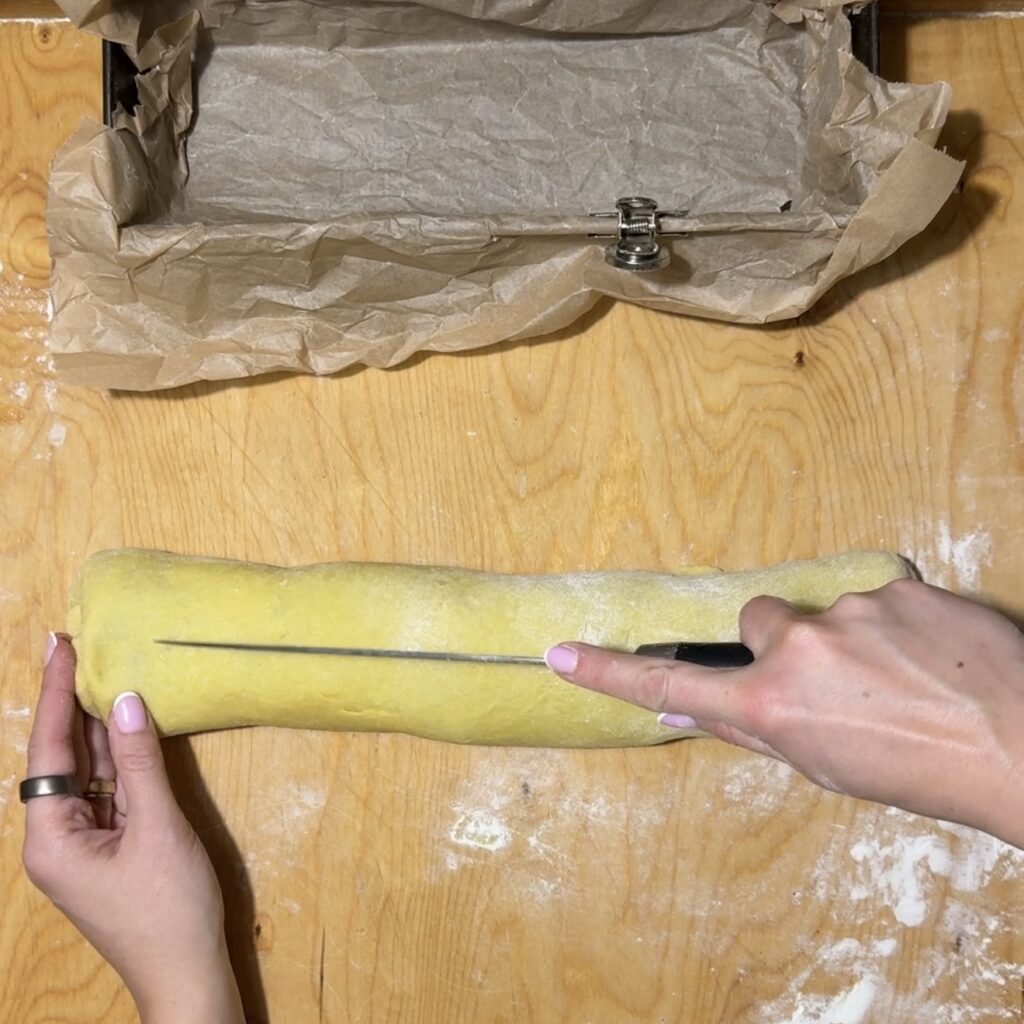 A person slices a rolled dough for Makowiec with a knife on a floured wooden surface; a parchment-lined baking tin sits nearby.