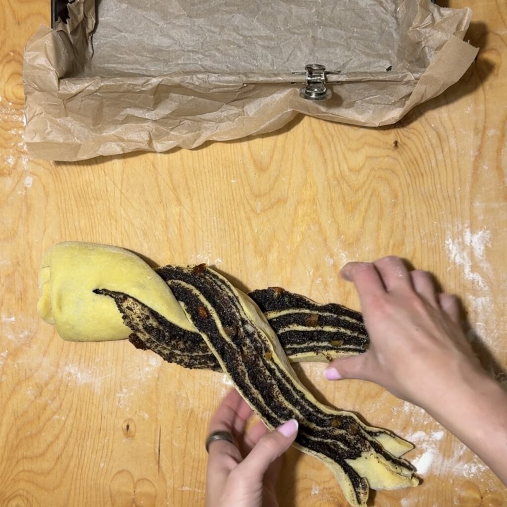 Hands twist a rolled dough with a dark poppyseed filling on a wooden surface, preparing Makowiec Zwijany, with a parchment-lined loaf pan visible at the top.