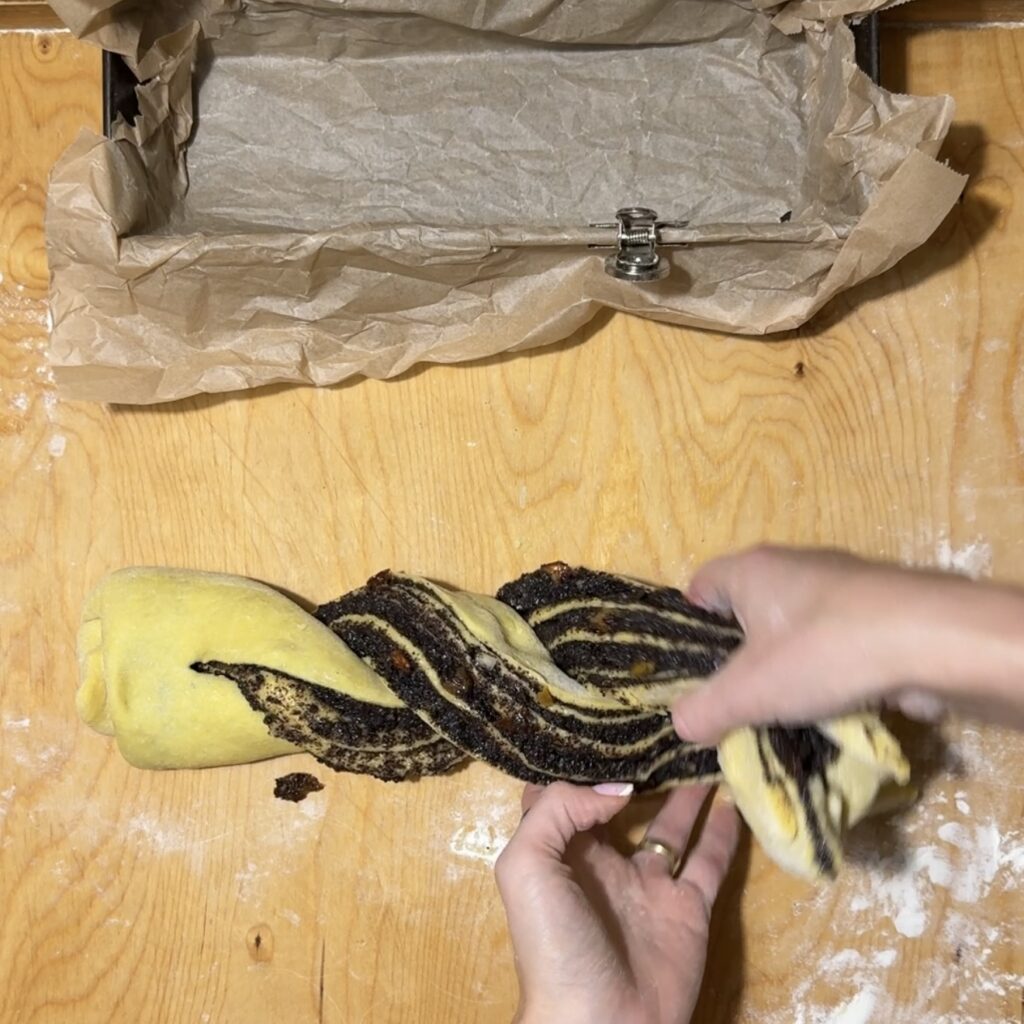 Hands twist a dough filled with chocolate spread, creating a twisted cake; a loaf pan lined with parchment paper is visible on the wooden surface.