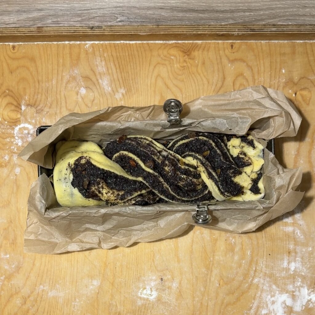 Unbaked poppyseed twisted cake dough, known as Makowiec Zwijany, is artfully twisted and placed in a parchment-lined loaf pan on a wooden surface.