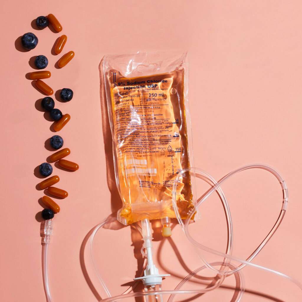 An IV therapy bag with a nutrient solution surrounded by pills and capsules against a peach-colored background in Meridian, Idaho.