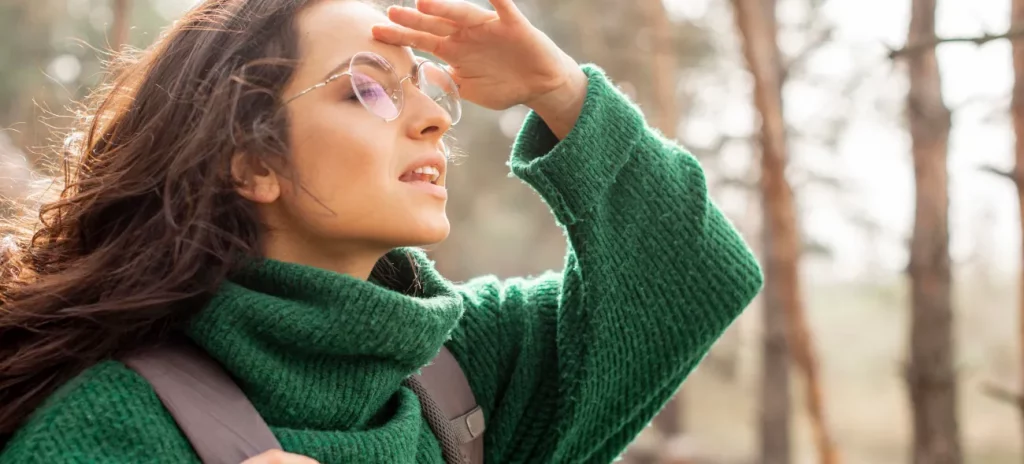 Auf der Suche nach dem eigenen Warum Eine junge Frau mit Brille und grünem Wollpullover ist auf der Suche nach dem Sinn des Lebens und blickt aufmerksam in die Ferne und hält eine Hand über die Stirn, um die Sicht zu verbessern. Im Hintergrund ist ein Wald zu sehen.