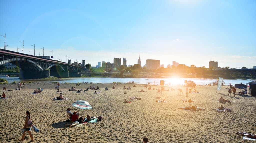 People relaxing on a sandy beach by a river with a bridge, city buildings, and a bright sun in the background&mdash;one of the lively beaches in Warsaw perfect for unwinding in the heart of the city.