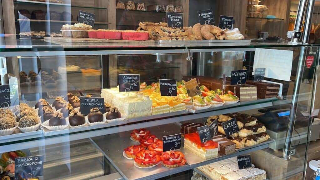 A glass display case filled with assorted cakes, pastries, and desserts, each labeled with small signs showing their names and prices&mdash;a tempting spot for the Best Breakfast Krak&oacute;w has to offer.