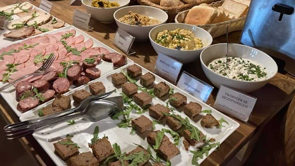 A buffet table with various cold meats, sliced bread, p&acirc;t&eacute;, and bowls of salad and cheese, each labeled with signs in Polish&mdash;perfect for enjoying the Best Breakfast Krak&oacute;w has to offer.