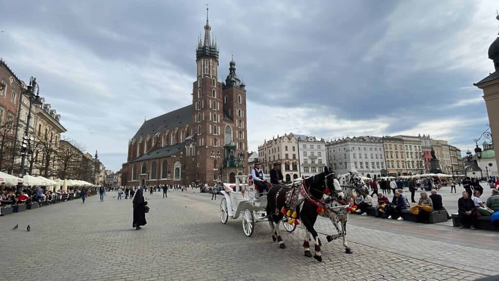 A horse-drawn carriage passes through a busy city square with St. Mary's Basilica and historic buildings in the background under a cloudy sky.