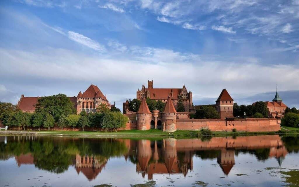 Large brick castle complex with towers and walls, surrounded by greenery and reflected in a calm body of water under a blue sky with scattered clouds&mdash;a true highlight among must-see castles for Poland tourism.
