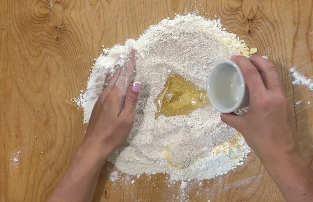 Hands preparing dough on a wooden surface, forming a well in a pile of flour with eggs, while pouring water into the center&mdash;capturing the first step to making Babcia's Pierogi.