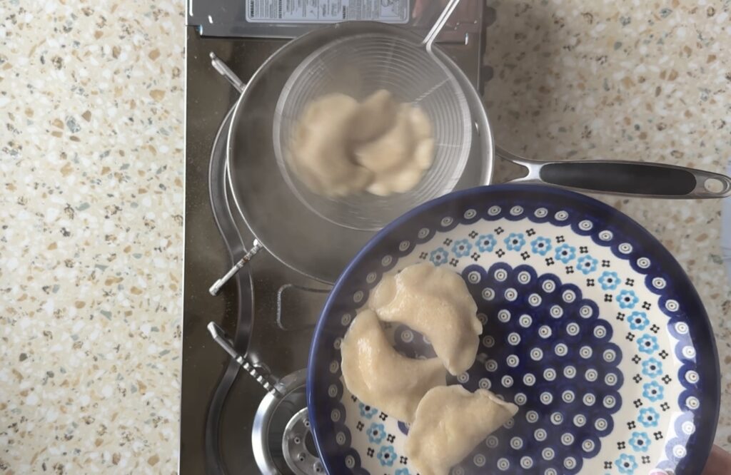 Three of Babcia's Pierogi are being lifted from a pot of boiling water with a slotted spoon, hovering above a patterned plate with three already-cooked dumplings.