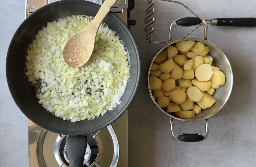 Chopped onions sizzle in a pan with a wooden spoon, beside a pot of peeled and halved potatoes and a potato masher&mdash;the classic start for Authentic Pierogi.