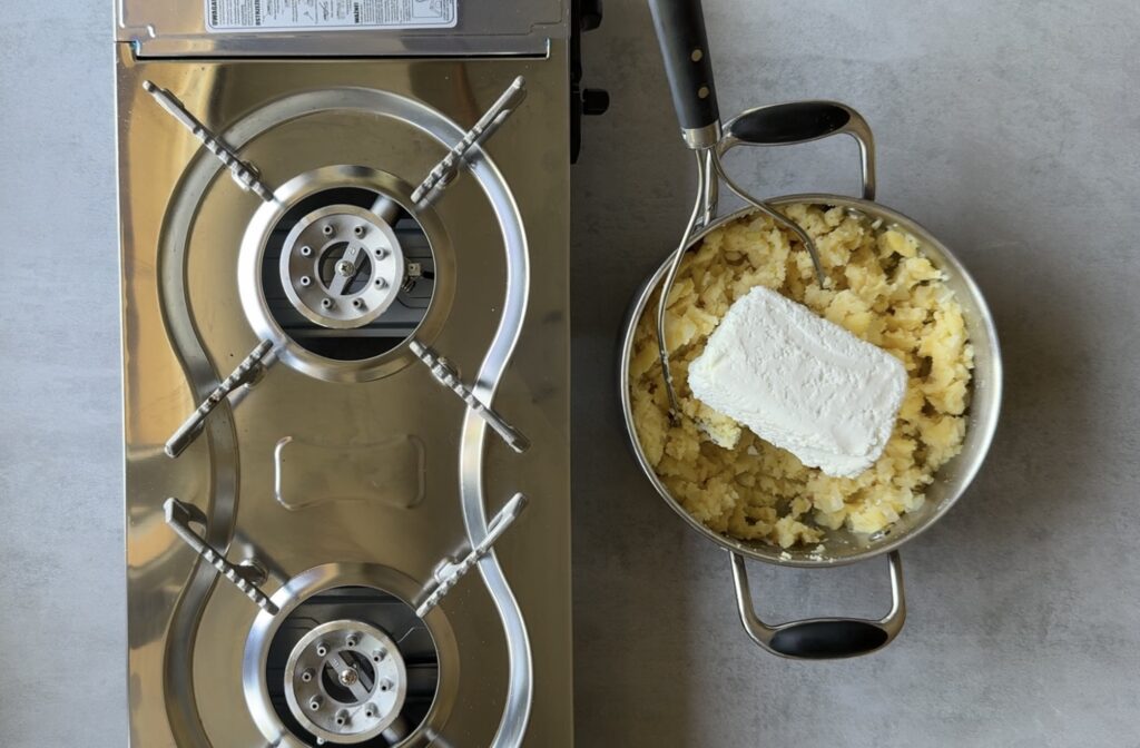 A metal pot with mashed potatoes and a block of cream cheese sits next to a stainless steel gas stove&mdash;perfect for crafting Authentic Pierogi just like Babcia's Pierogi recipes.