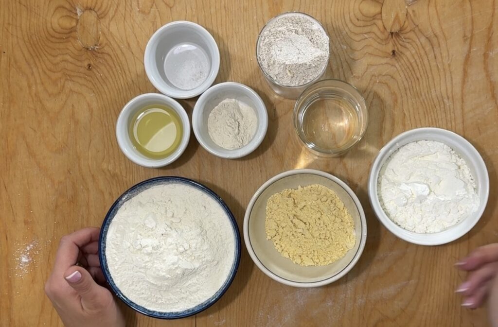 A wooden table with nine bowls containing baking ingredients for Authentic Pierogi, including various flours, oil, water, and salt. Two hands hold a bowl of all-purpose flour in the lower left corner.