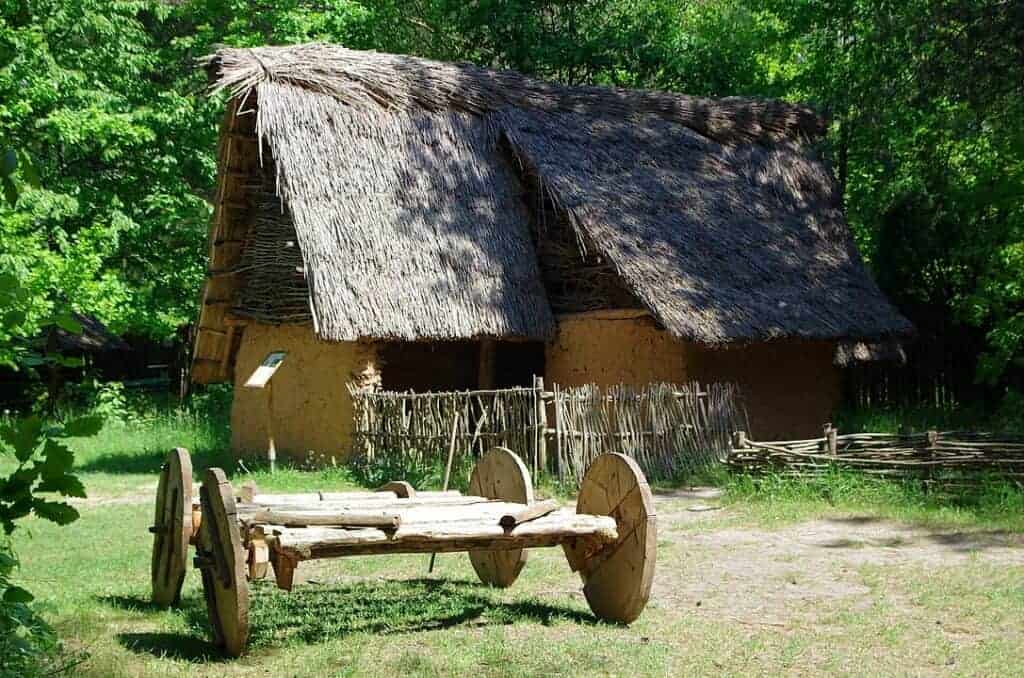 A rustic wooden cart stands in front of a traditional thatched-roof hut in Swietokrzyskie, one of Poland’s charming places to visit, surrounded by trees and simple wooden fences on a sunny day.