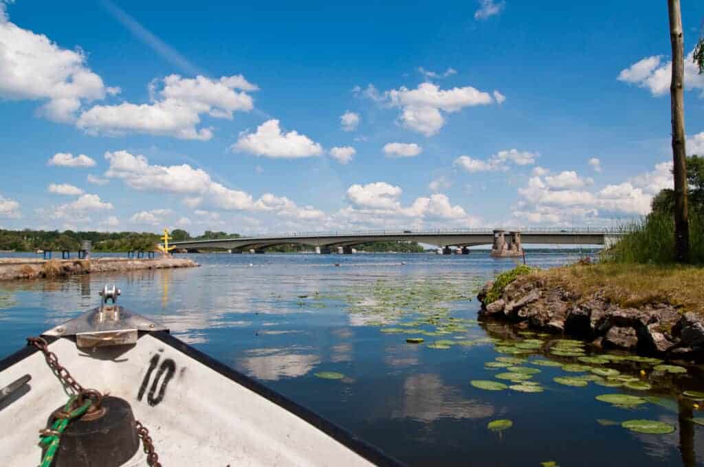 View from a boat on a calm river with lily pads, looking toward a concrete bridge under a blue sky with scattered clouds&mdash;just moments away from some of the best beaches in Warsaw.