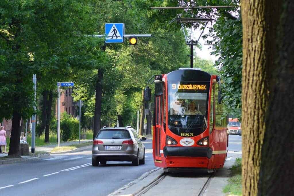 A red tram labeled "9 Chorz&oacute;w Rynek" travels along tracks beside a road with a car and a pedestrian crossing sign overhead, surrounded by trees&mdash;a charming scene in Slaskie Poland, one of the Best Places Slaskie to explore.