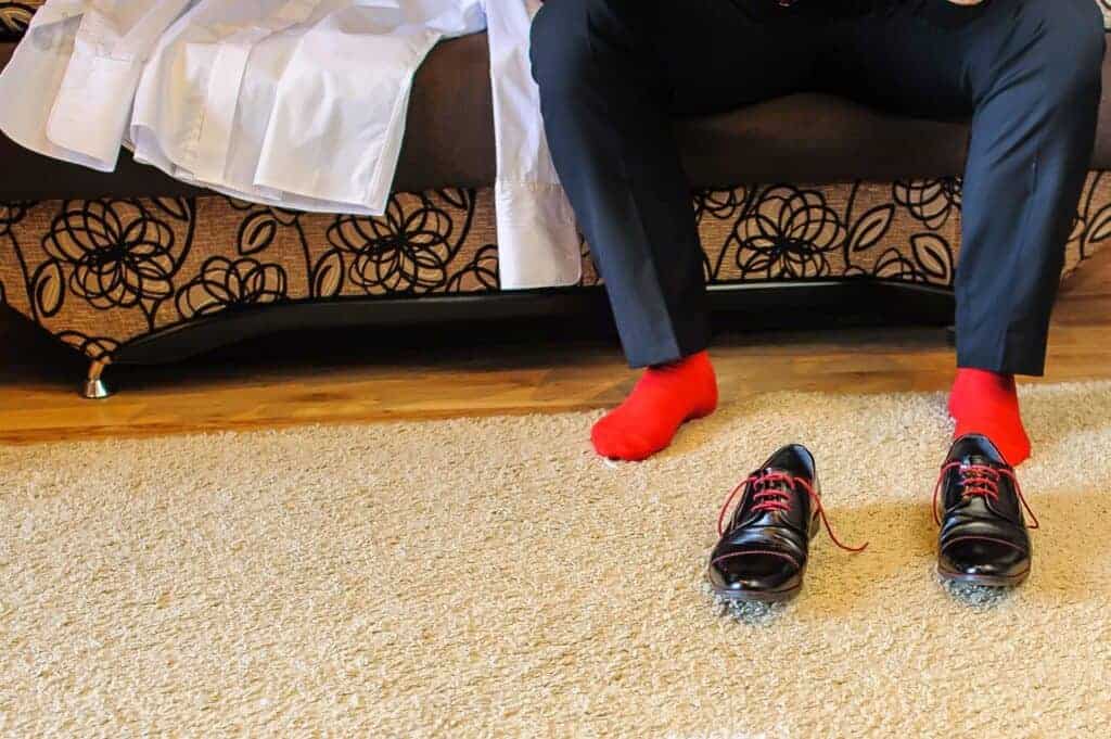 A person in red socks and black pants sits on a bed, their black dress shoes with red laces resting on a beige carpet&mdash;perhaps pondering Polish etiquette or things to avoid in Poland before their journey.