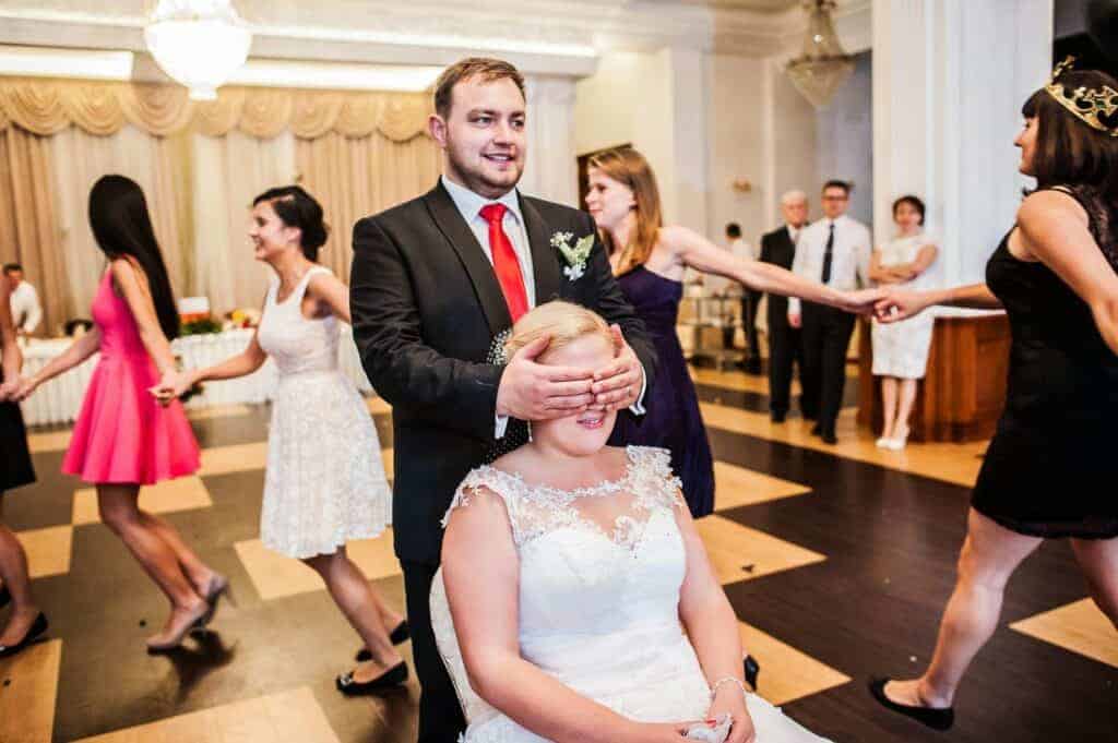 A man in a suit covers the eyes of a seated woman in a white dress&mdash;a charming moment inspired by Polish wedding traditions&mdash;while people hold hands and dance in a decorated hall.