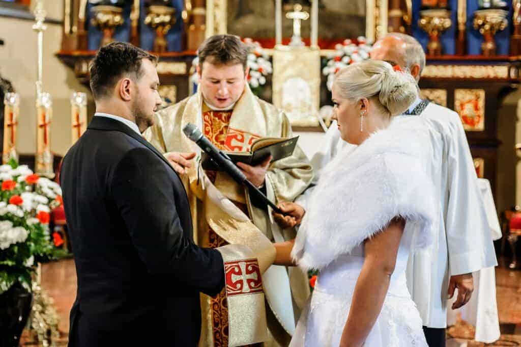 A bride and groom stand before two priests during a church wedding ceremony, holding hands as a priest reads from a book and another holds a microphone, reflecting cherished Polish wedding traditions.