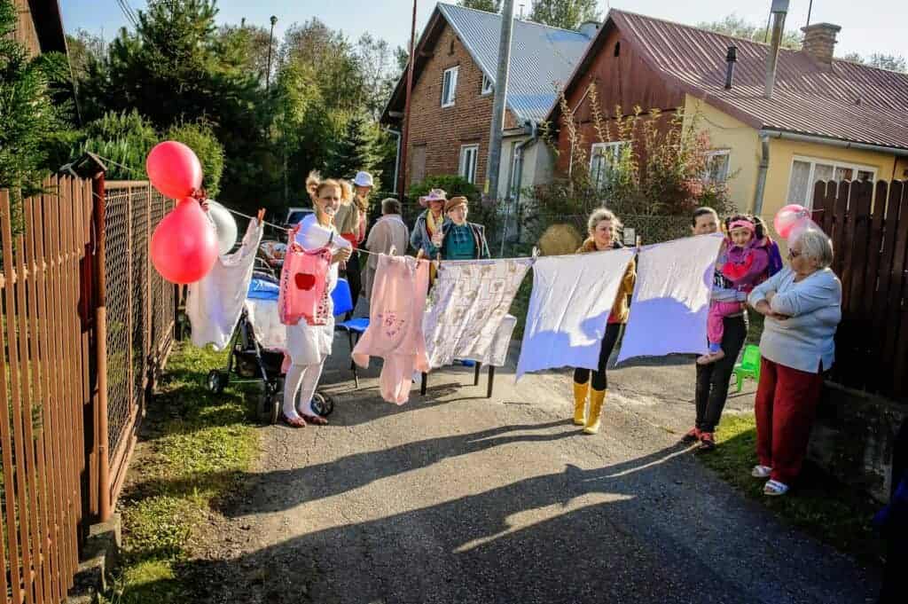 A group of people stand outside houses, holding a clothesline with laundry and pink balloons attached, celebrating in the spirit of Polish wedding traditions on a sunny day.