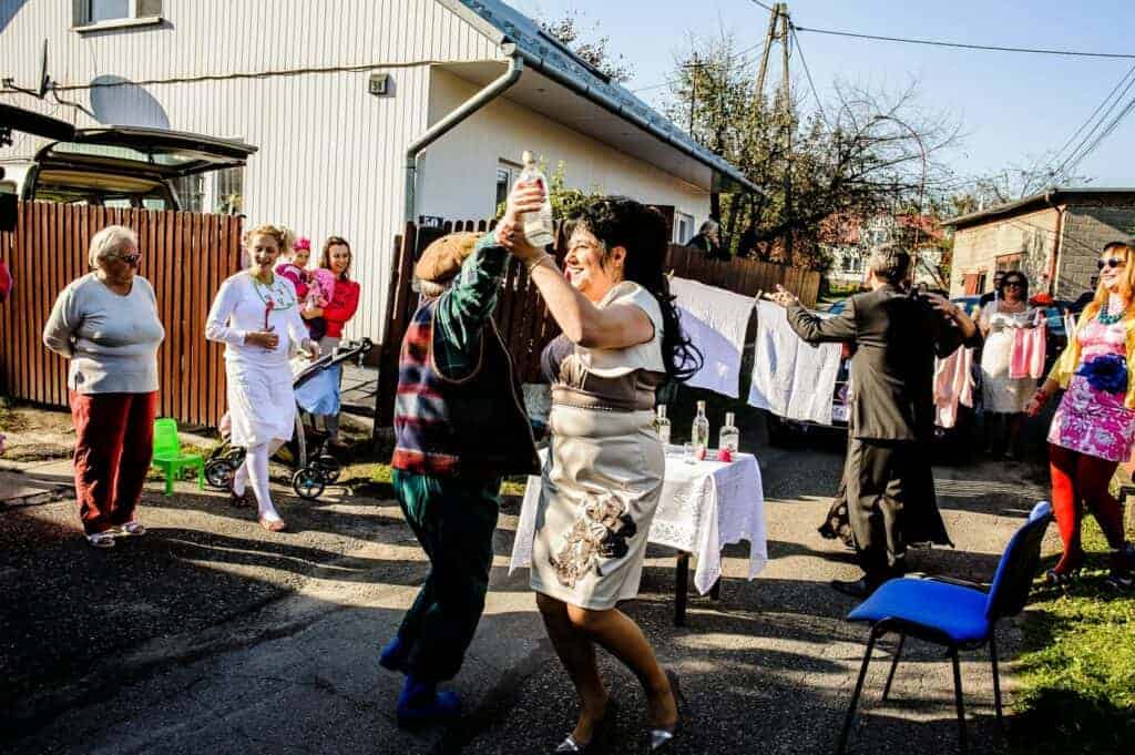People dance and socialize outdoors near a table with drinks, while others stand and talk in a residential area with laundry hanging nearby, capturing the lively spirit of Polish wedding traditions.