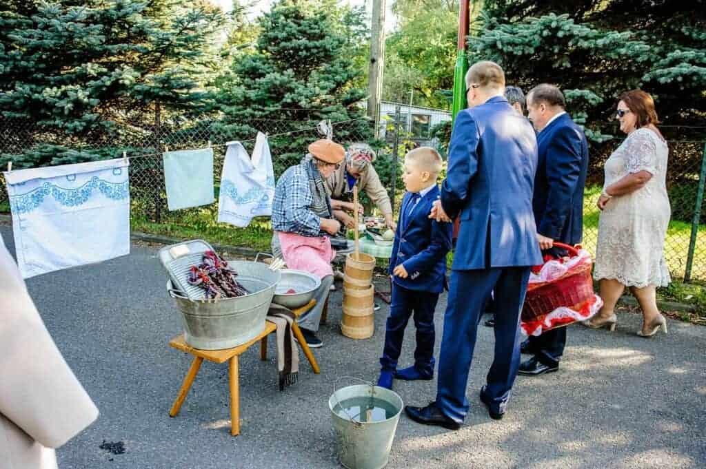 People in formal attire watch two individuals demonstrating traditional laundry washing outdoors&mdash;a nod to Polish wedding traditions&mdash;with linens hanging on a fence and pine trees in the background.