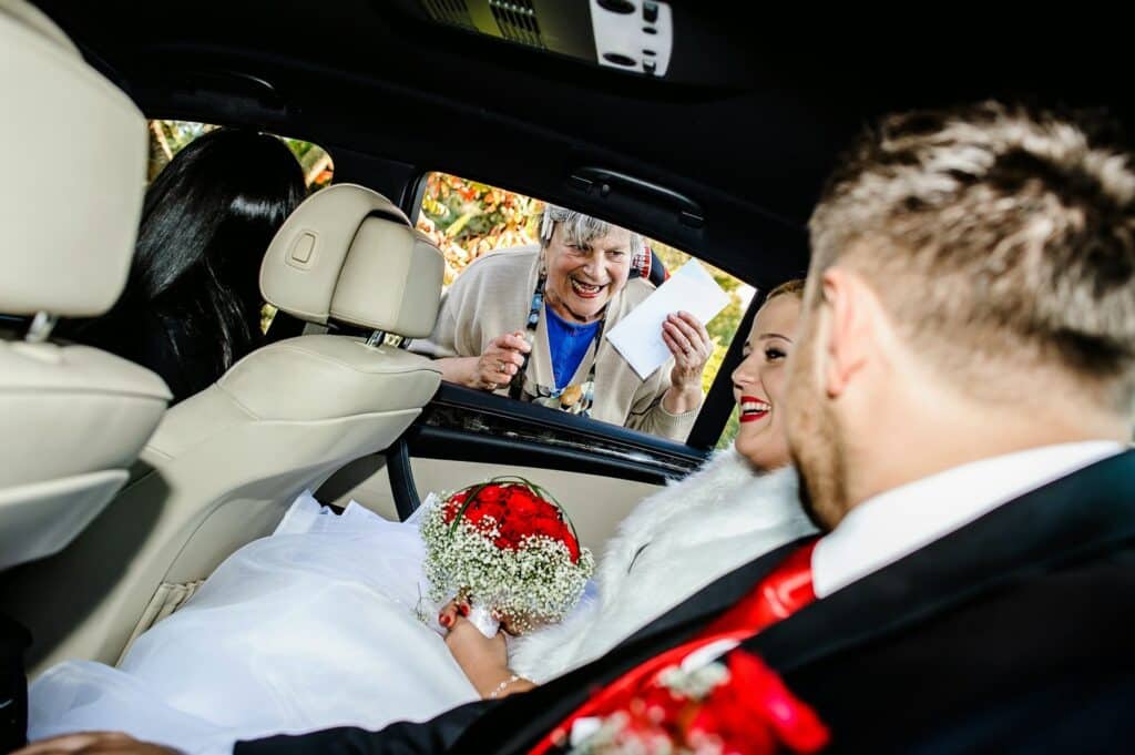 A bride and groom sit in a car, smiling at an older woman standing outside the window who is holding an envelope&mdash;a joyful moment reflecting Polish wedding traditions.