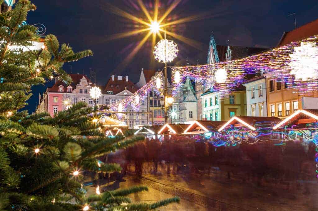 A decorated Christmas tree and festive lights overlook a crowded outdoor market with colorful buildings at night, capturing the magical spirit found at the Best Christmas Markets in Poland.