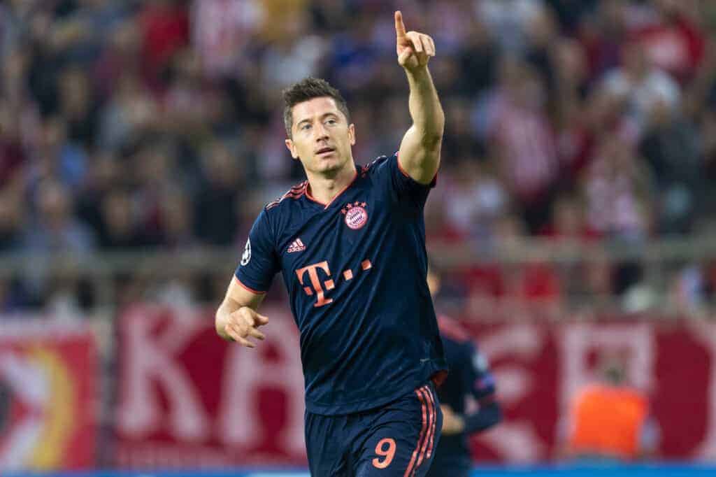 A soccer player born in Poland in a dark Bayern Munich jersey with number 9 raises his right arm and points upward on the field during a match, highlighting one of the most famous Polish celebrities in football.