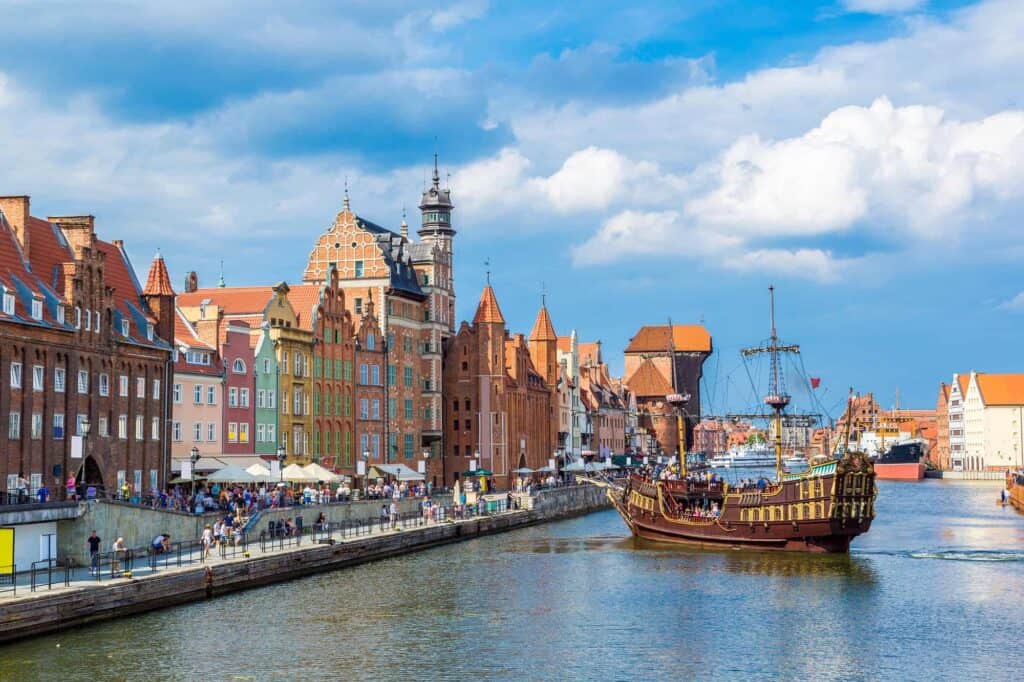 A replica pirate ship sails along a river bordered by colorful historic buildings and a lively promenade in Gdańsk, Poland, under a partly cloudy sky.