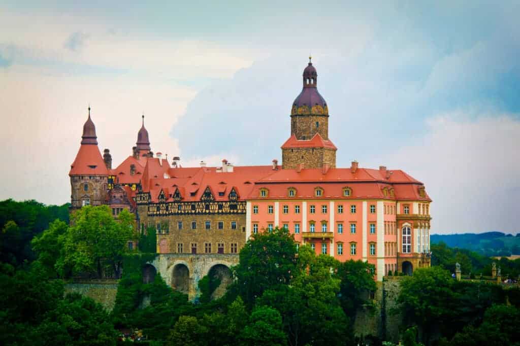 A large castle with red roofs and multiple towers sits atop a hill surrounded by trees under a cloudy sky&mdash;one of the must-see castles that stands out among Poland attractions.
