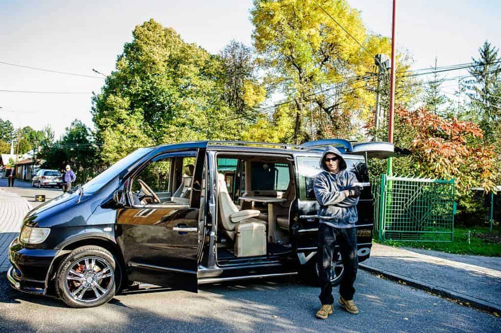 A person in a dark hoodie stands with crossed arms next to a black van with open doors on a sunny day, as if guarding it&mdash;a scene reminiscent of secretive moments sometimes seen at Polish weddings. Trees and a green fence are visible in the background.