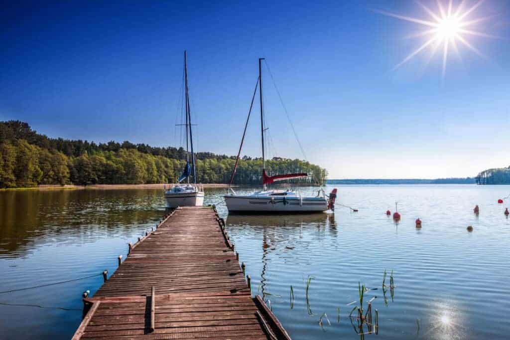 A wooden dock extends into a calm lake with two sailboats moored beside it under a clear blue sky and bright sun, evoking the serene beauty found near many historical landmarks in Poland. Trees line the distant shore.