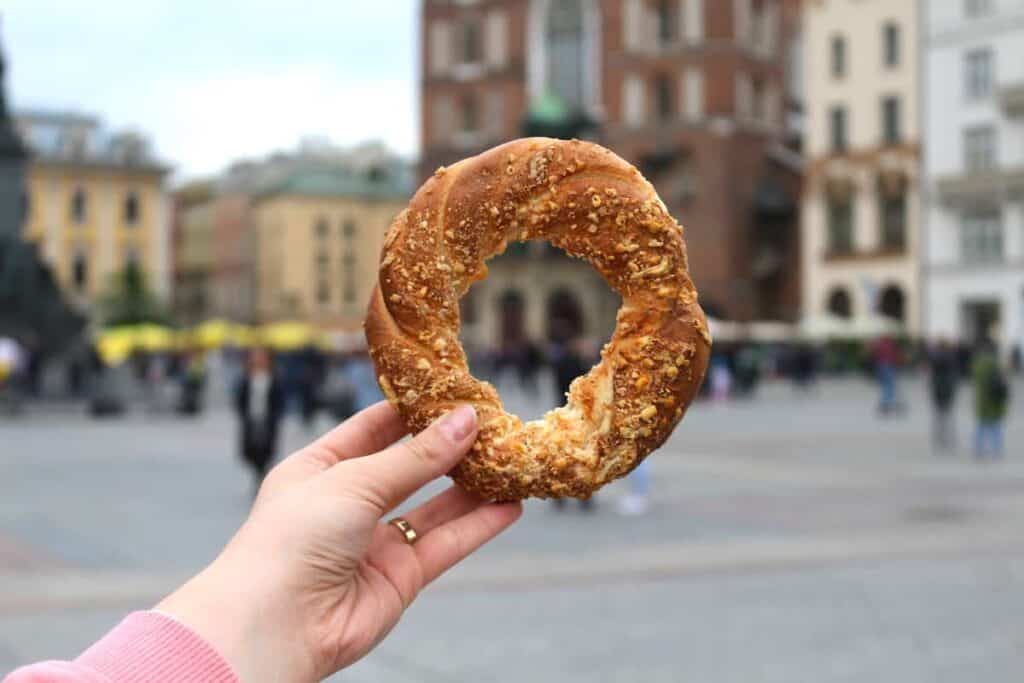 A hand holding a round, sesame-topped bread with a bite taken out, in focus against a blurred city square background.