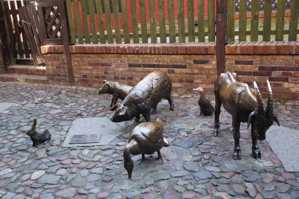 Bronze statues of a duck, goose, pig, goat, heron, and rooster are arranged on a cobblestone walkway in front of a brick and wooden fence&mdash;an example of charming Wroclaw Street Art.