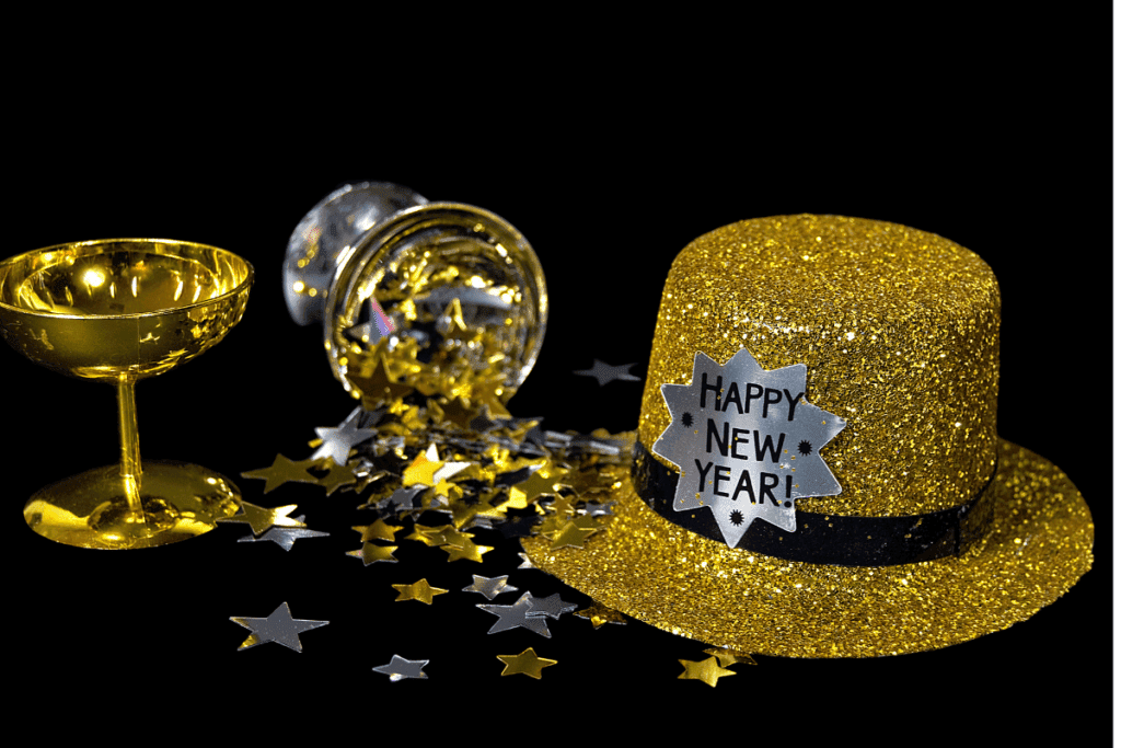 A gold glittery "Happy New Year!" hat sits beside two overturned golden glasses and scattered metallic star confetti on a black background, evoking the festive spirit of Polish New Year's traditions.