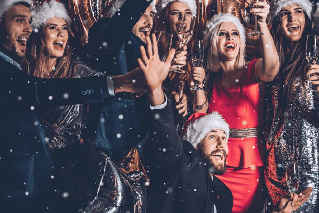 A group of people in festive outfits and Santa hats celebrate together indoors, holding champagne glasses and smiling amid falling confetti, embracing Polish New Year's traditions.