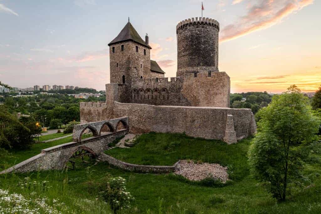 A medieval stone castle with a round tower and arched bridge sits on a grassy hill at sunset, surrounded by trees and distant buildings&mdash;a true gem among must-see castles in Poland.