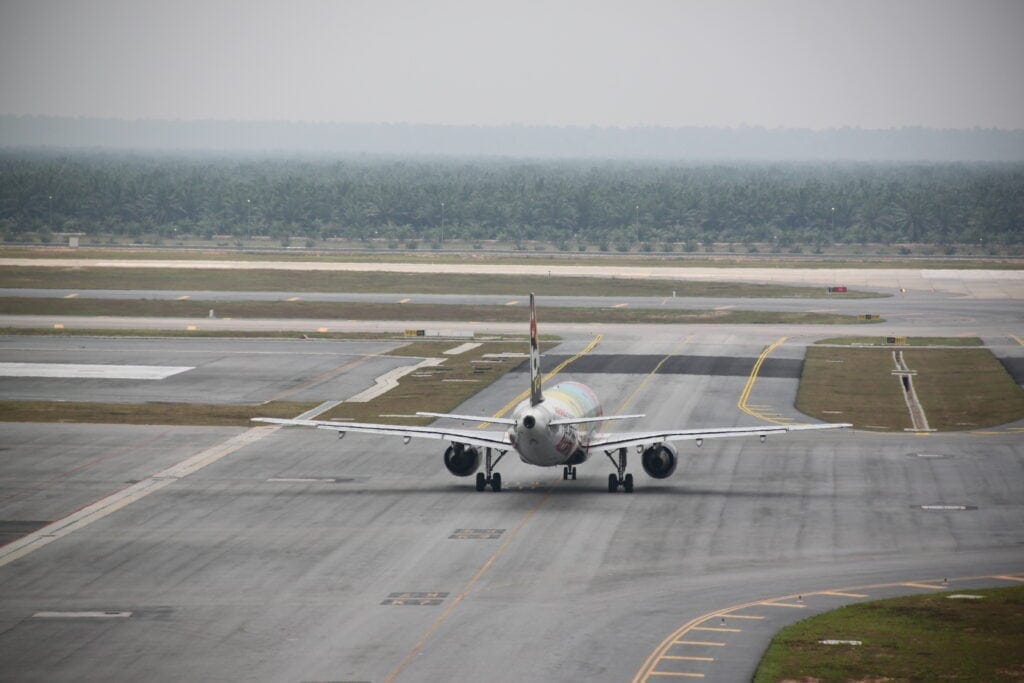 A commercial airplane taxis on a runway at an airport under overcast skies, with a line of trees in the background, ready for its flight to Poland.