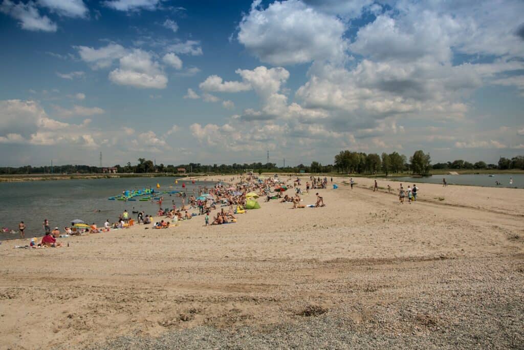 A sandy beach with many people sunbathing, swimming, and relaxing near the water under a partly cloudy sky&mdash;one of the lively Krakow beaches perfect for a summer day.