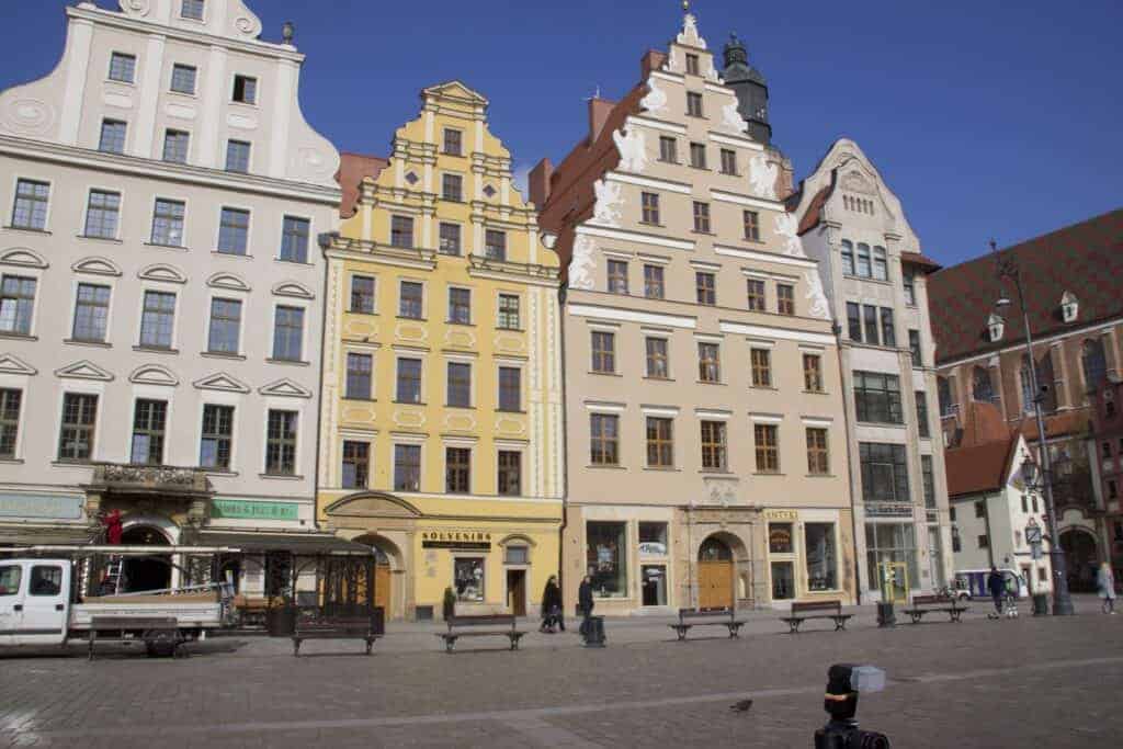 Row of colorful, historic buildings with ornate facades and gabled roofs face a cobblestone square in Wroclaw, known for its travel safety. Benches, a few people, and a pigeon add charm to this safe and welcoming city scene.