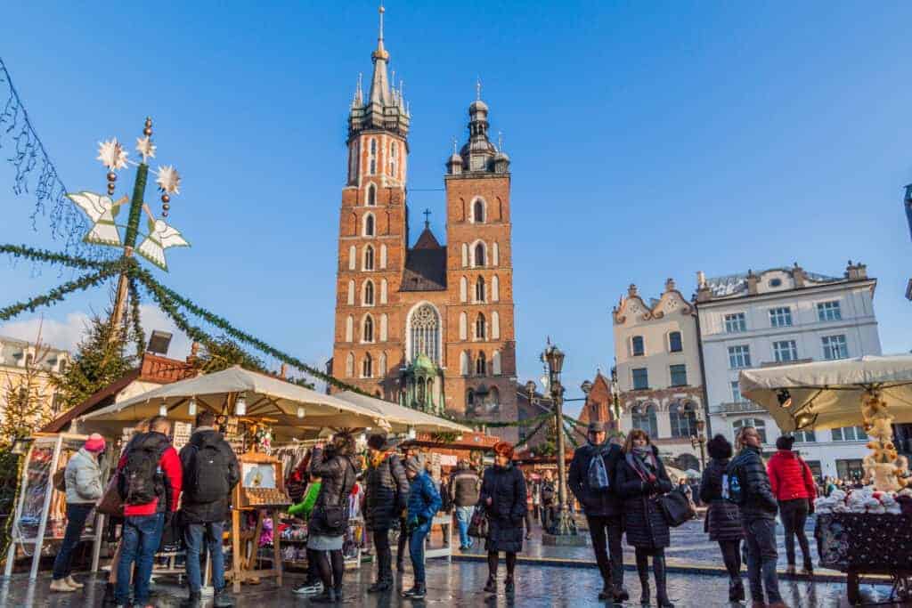 People walk through an outdoor market with festive stalls in front of St. Mary&rsquo;s Basilica on a sunny day, capturing the charm of Christmas Markets Poland in a historic European city square.