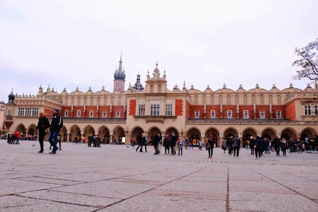 People walk across a large open square in front of the historic Cloth Hall building in Krak&oacute;w, Poland, a must-see spot for anyone planning a 3 days itinerary. Towers rise impressively in the background.