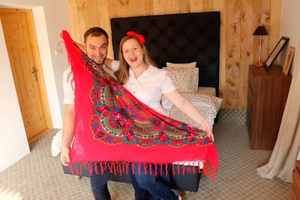 A smiling man and woman stand in a bedroom holding a large red patterned shawl, one of their cherished traditional Polish gifts, spread out in front of them.