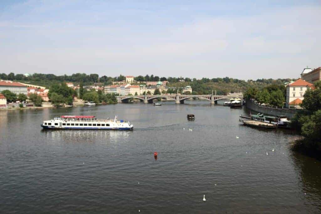 A river cruise boat travels along a wide river with a bridge, greenery, and buildings in the background under a partly cloudy sky&mdash;one of the best options for unique travel options between Krakow to Prague.