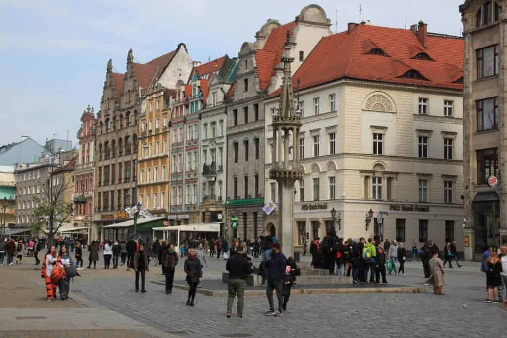 People walk and gather in a cobblestone square lined with colorful historic buildings featuring ornate architectural details and red roofs, creating a vibrant atmosphere that reflects Wroclaw&rsquo;s safety and welcoming charm.
