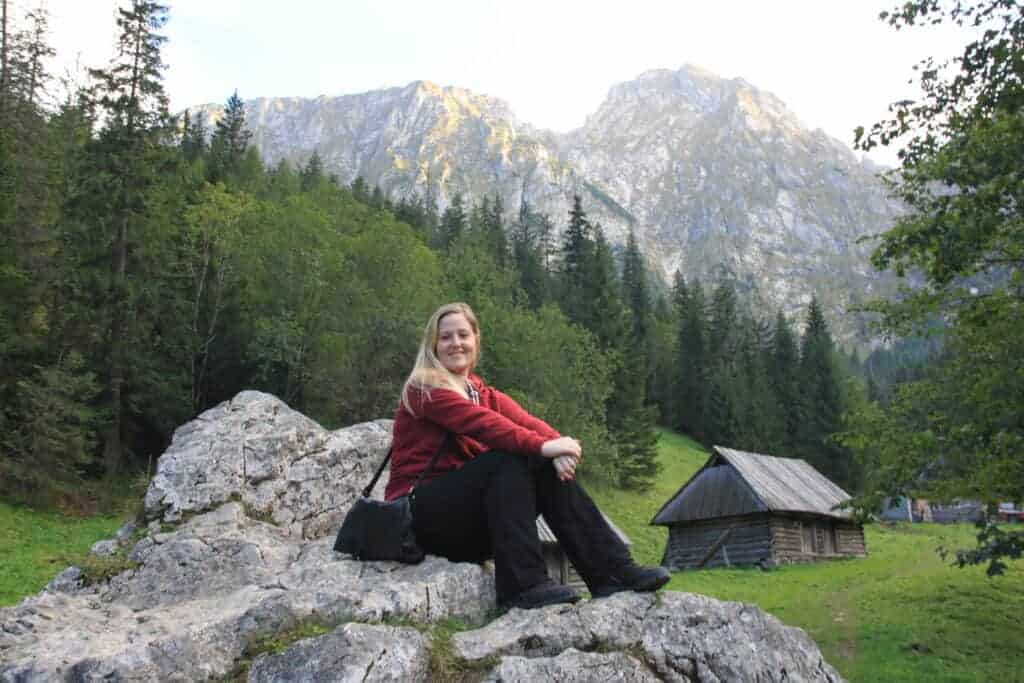 Woman in a red jacket sits on a large rock, smiling, with trees, wooden cabins, and mountains in the background—perfect inspiration for solo travel Poland adventures.