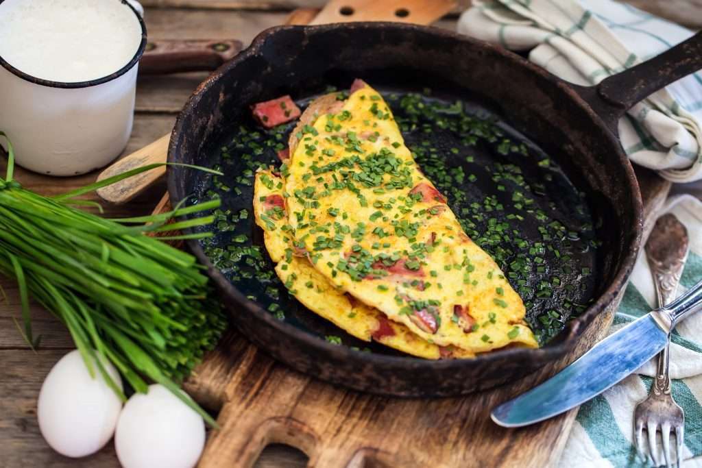 A freshly cooked omelette with herbs and diced Idaho potato in a cast iron skillet, accompanied by kitchen utensils and raw ingredients on a wooden table.
