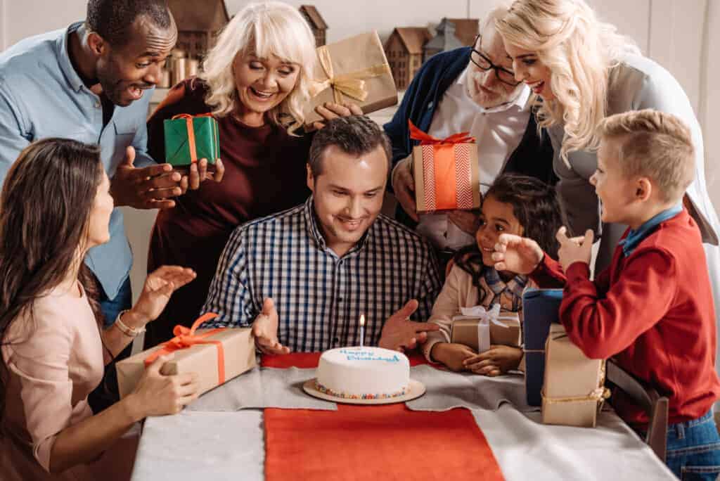A group of people gathers around a man at a table with a cake, smiling and holding wrapped gifts, enjoying a joyful Name Day celebration.